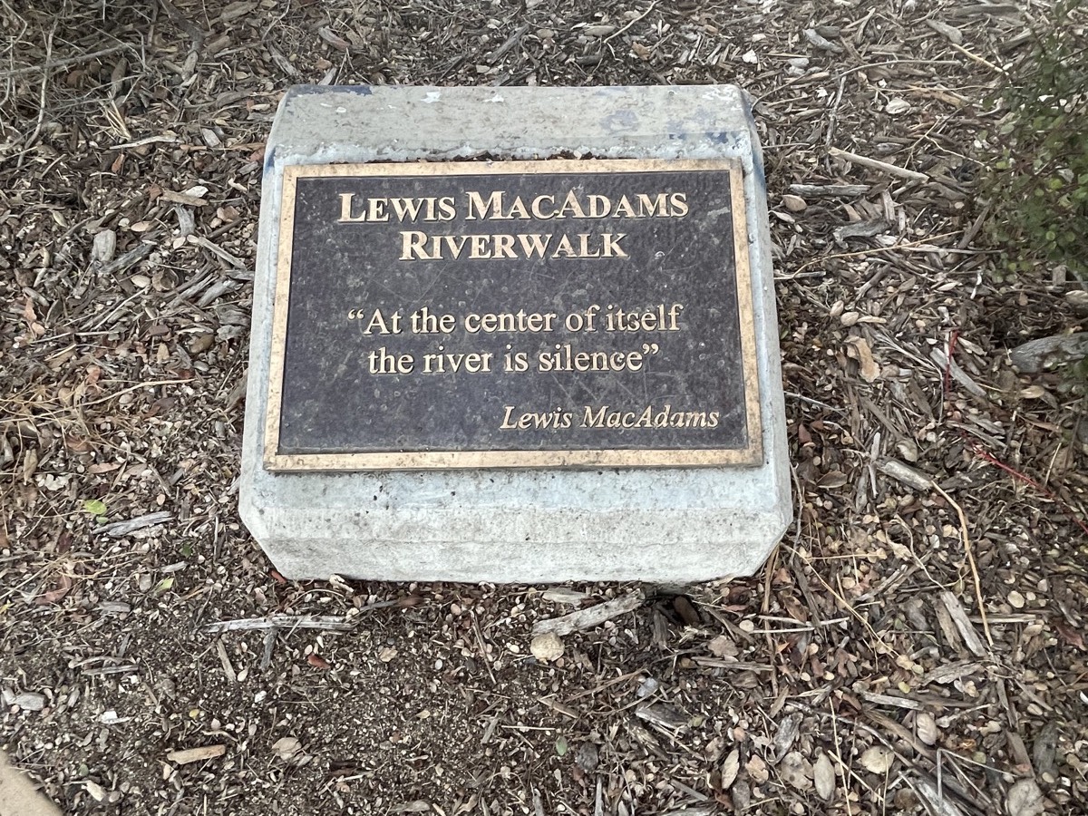 A plaque affixed to a stone at ground level. It reads “Lewis MacAdams Riverwalk. At the center of itself the river is silence.”